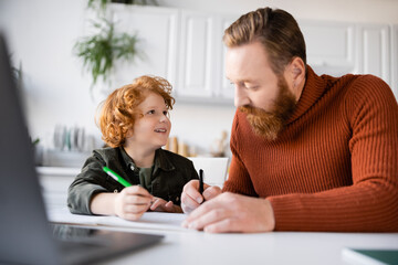 Fototapeta premium happy boy looking at bearded father while doing homework near blurred laptop