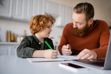 Fototapeta premium bearded man talking to redhead son doing homework near blurred laptop