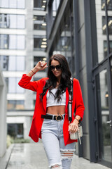 Stylish casual pretty woman in fashionable urban outfit with a red blazer, jeans and a white top with a purse walking in the city and wearing sunglasses