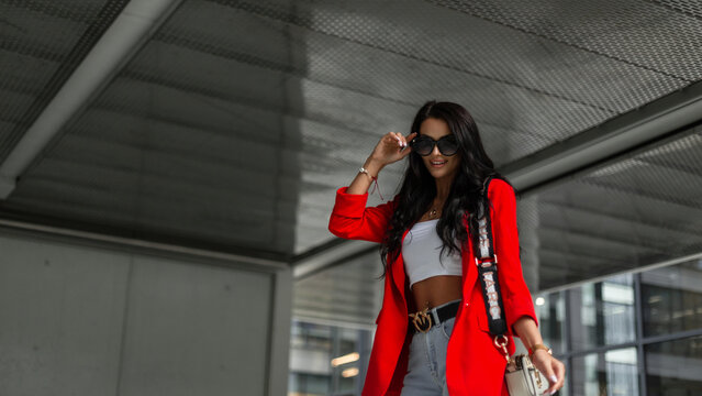 Fashionable beautiful happy happy successful business girl in fashion bright clothes with a red blazer, top and jeans with a purse wears sunglasses and walks in the city