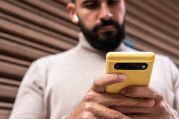 Bearded young man in turtleneck sweater with wireless earphones holding his yellow phone with both hands typing text messages.