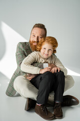 Smiling man in stylish jacket embracing red haired kid on grey background with sunlight