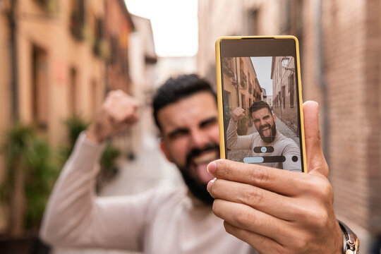 Young man with a beard wearing a turtleneck taking a selfie raising an arm in a sign of strength and sticking out his tongue in one of the most prominent streets of his city.