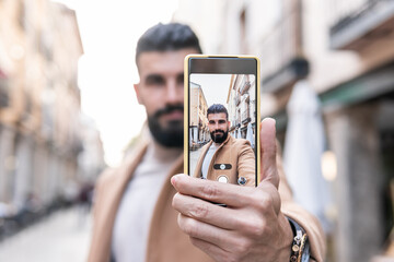 Young man with a beard, turtleneck and jacket taking a self photo with his cell phone in the city center.