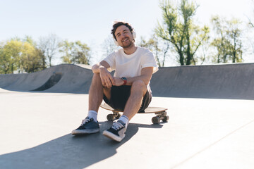 Smiling skater sitting on longboard during break listening to music on headphones