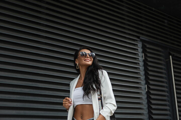 Beautiful young happy girl with a smile with fashion sunglasses in stylish white clothes with a blazer and a top walks in the city near a black metal wall. Urban female summer style look clothes