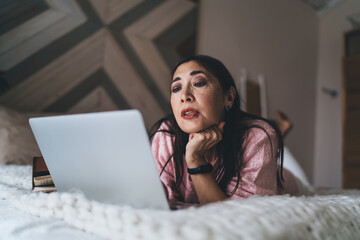 Thoughtful mature ethnic woman using laptop on bed