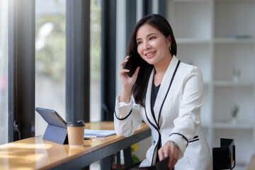 Asian businesswoman sitting with her laptop happily chatting on the phone with a customer explaining in detail about her work and company products in the office.