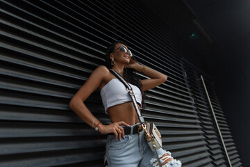 Fashionable beautiful stylish woman model with a smile with fashion sunglasses in summer trendy clothes with a top, ripped jeans and a small bag near a black metal wall in the city