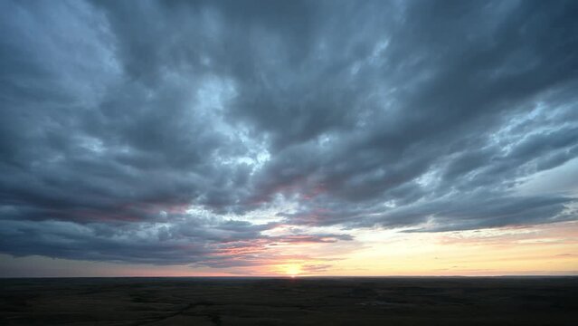 Time lapse of a sunset over prairie badlands with teal blue to orage colored clouds moving towards the viewer.
