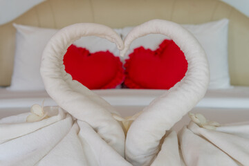Close-up of a set of white towels curled into a swan-shaped heart on the double bed in front of two red heart-shaped cushions.