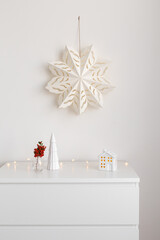 Christmas cozy winter home decor. New year interior decorations. Paper snowflake on wall, branch with red berries in vase, decorative ceramic house and christmas tree. Stylish composition on dresser.