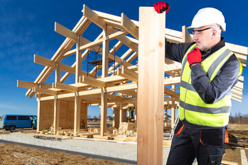 Fototapeta premium Construction company worker. Man in construction uniform thought. Man builder scratches chin. Guy holds beam for building house. Foreman stands at construction site. Construction country cottage