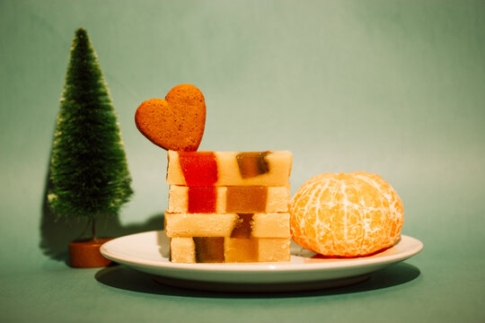 Marzipan marmalade turrone on plate, tangerine on green emerald background. Spanish dessert made from almonds, honey, sugar, eggs. Christmas sweets, heart-shaped cookie. New Year pine tree decor 2023.