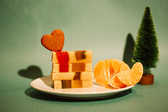 Marzipan marmalade turrone on plate, tangerine on green emerald background. Spanish dessert made from almonds, honey, sugar, eggs. Christmas sweets, heart-shaped cookie. New Year pine tree decor 2023.