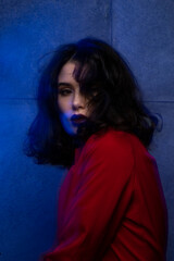 Beauty portrait of young woman with makeup and hairstyle, wear red suit, looking at camera sensual, over blue neon studio light.
