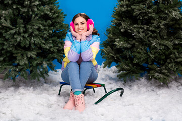 Photo of adorable charming lady wear striped coat sitting sledge between xmas fir indoors isolated blue color background