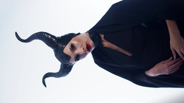 Beautiful caucasian woman in a maleficent costume. A woman seduces with hand gestures and a mystical look. A girl in a black devilish costume with horns on a white background. Vertical video.