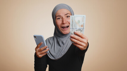 Happy muslim woman in hijab looking smartphone display sincerely rejoicing win, receiving money dollar cash banknotes, success lottery luck. Young millennial girl isolated on beige studio background