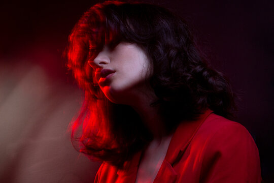 Fashion photo of young woman with makeup and hairstyle, wear red suit, with closed eyes, red neon studio light.