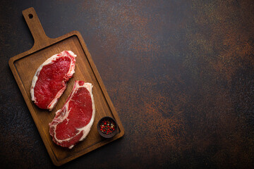 Fototapeta na wymiar Two raw uncooked meat beef rib eye marbled steaks on wooden cutting board with seasonings on dark rustic background ready to be grilled from above, preparing dinner with meat, space for text