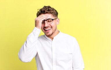 young adult caucasian man laughing and slapping forehead like saying d’oh! I forgot or that was a...