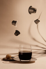 Levitating coffee capsules with glass of black coffee on paper background with leaf shadow front view