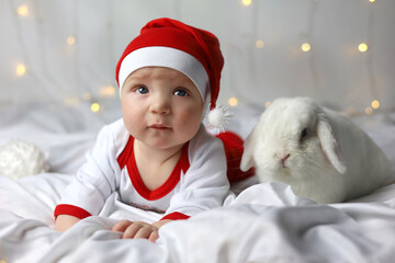 Baby and white rabbit lie in bed. Happy litlle kid in red Christmas hat with rabbit. Child's Christmas gift in bed. Boy in a Santa hat with a rabbit. Funny pet. Christmas lights are on