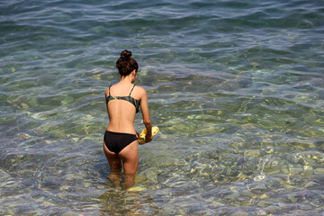 Girl in black swimsuit washes swimming shoes in sea water. Vacation on a pebble beach of sunny resort