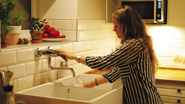 Concept of pure drinking water. Woman pouring clean filtered water from the tap in glass for drinking at modern loft style kitchen at night or evening. Kitchen with lights. Thirsty young female 