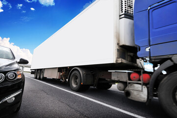 Close-up of a blue truck with a white trailer on a highway with other car against a sky with clouds