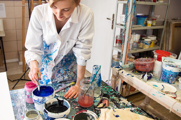 Portrait of young woman artist in robe covered with various stains stirring blue acrylic paint in bucket with brush.