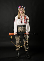 Full length portrait of a Ukrainian female soldier wearing a Ukrainian embroided shirt with a wreath and holding a rifle isolated on a black - 546998187