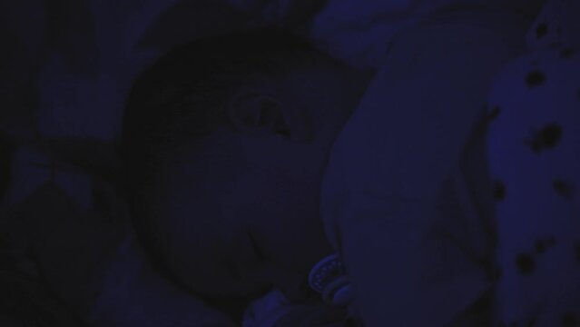 Cute child sleeps in crib at night. Warm light from twinkling lights falls on bed. Baby boy sleeping in crib in evening in light of night twinkling lights. 