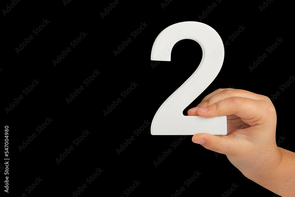 Poster close-up hand holding a white number 2 on a black background. number two isolate on black background