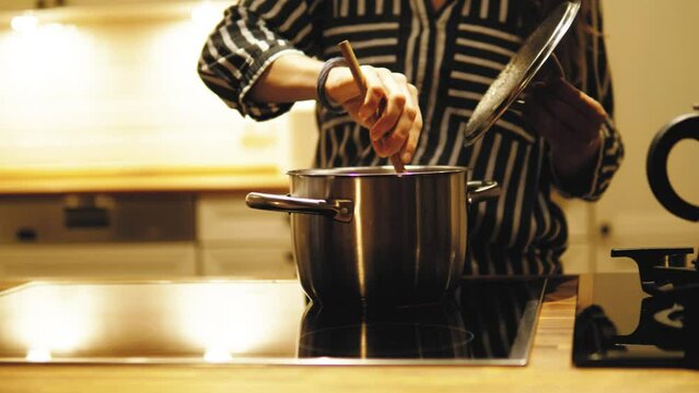Young woman shef cooking, mixing ingredients in the pot with wooden spoon. Housewife woman cooking dish or soup in the pot, standing in the kitchen at night or evening. Delicious dish. Preparation