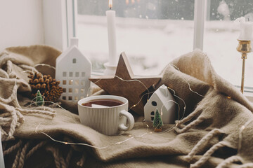 Winter hygge. Warm cup of tea, candles, lights, little christmas houses and trees, wooden star on...
