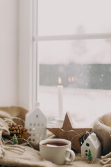 Cozy home at snowy day. Warm cup of tea, candles, lights, little christmas houses and trees, wooden star on cozy blanket on windowsill. Winter hygge. Atmospheric scandinavian mood