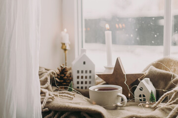 Winter hygge. Warm cup of tea, candles, lights, little christmas houses and trees, wooden star on cozy blanket on windowsill. Cozy home. Atmospheric snowy day. Christmas background