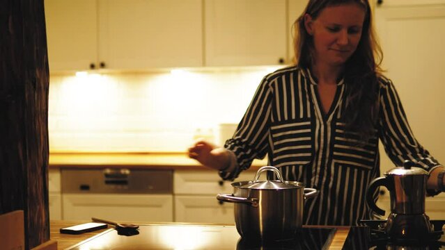 Female cooking in the kitchen, looking for recipe on smartphone, standing near oven and simmering water in the pot, mixing putting salt in it in the evening or late night. Cozy modern kitchen at night