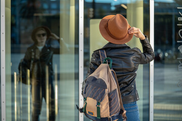 30s woman with short blonde hair in leather jacket and trendy hat looking on her reflection in mirror building in Barcelona city