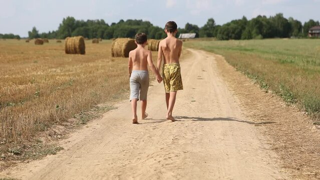 Two children-brothers without t-shirts and without footwear wolking from the camera holding hands on a sandy rural village road in a sunny day. There are haystacks near. 4k
