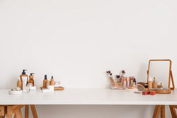 Different cosmetics and makeup accessories on table against light wall