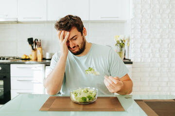 A sad young man looks longingly at a salad, holds a fork with greens in his hand, and a plate of...