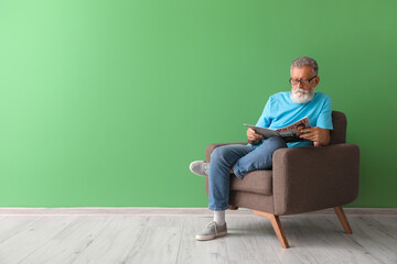 Senior man reading magazine in armchair near green wall