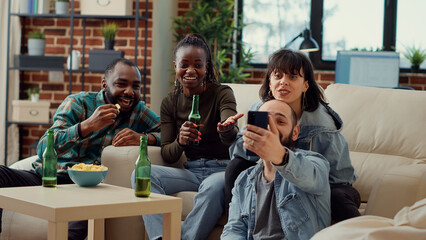 Diverse people watching funny video on smartphone, drinking beer from bottles and having fun at home gathering. Group of friends enjoying online internet on mobile phone. Handheld shot.
