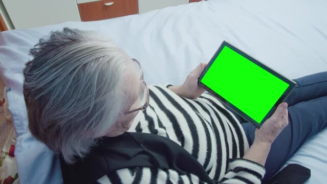 Old grey-haired woman in striped sweater and eyeglasses reads electronic book using green screen tablet PC and lying on wooden bed upper view