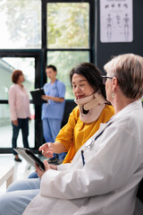 Senior medic consulting patient with cervical neck collar using tablet to take notes, doing checkup visit in waiting room lobby. Asian woman with physical pain and injury having medical appointment.