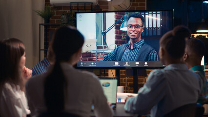 Remote team leader listening to employees report in videocall, diverse people talking in videoconference at night time. Office workers chatting in teleconference, executive speaking. Tripod shot.