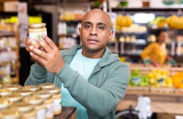 Interested Latin American man choosing foods in supermarket, reading jar contents on label